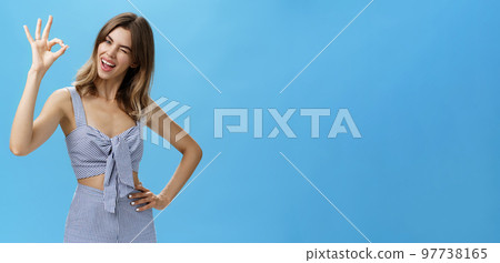 Cute charismatic woman with gap teeth showing okay gesture tilting head joyfully, holding hand on waist and winking with self-assured expression reassuring friend everything goes on plan Cute charismatic woman with gap teeth showing okay gesture tilting head joyfully, holding hand on waist and winking with self-assured expression reassuring friend everything goes on plan 97738165