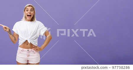 Thrilled and excited positive attractive woman with fair hair tanned skin and tattooed arms smiling joyfully with opened mouth staring impressed and delighted at camera, pointing left over purple wall 97738286