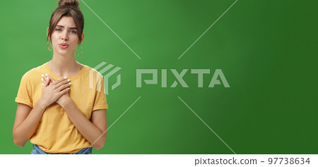 Touched and pleased attractive young woman in yellow t-shirt holding palms pressed to chest in thankful and grateful gesture folding lips melting heart seeing cute thing over green background 97738634