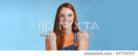 Indoor shot charismatic assertive happy smiling redhead woman orange shirt denim overalls pointing sideways thumbs left right showing choices opportunities give chance choose, blue background 97738707
