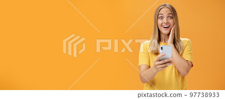 Portrait of surprised and impressed woman reacting to awesome app in smartphone touching cheek from amazement and joy smiling broadly at camera holding cellphone in hand over orange wall Portrait of surprised and impressed woman reacting to awesome app in smartphone touching cheek from amazement and joy smiling broadly at camera holding cellphone in hand over orange wall 97738933