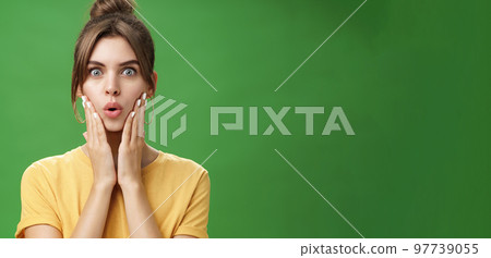 Portrait of impressed and shocked cute feminine woman in yellow t-shirt folding lips from excitement and interest touching cheeks surprised reacting to amazing rumor posing over green background Portrait of impressed and shocked cute feminine woman in yellow t-shirt folding lips from excitement and interest touching cheeks surprised reacting to amazing rumor posing over green background 97739055