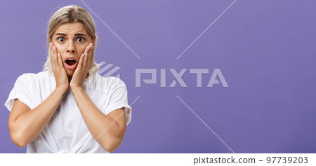 Shocked and worried silly european woman expressing empathy feeling sorry for poor animal in danger gasping opening mouth holding hands on face frowning feeling troubled over purple wall 97739203