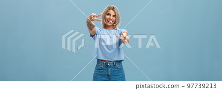 Beautiful tattooed girl enjoying weekends sticking out tongue joyfully winking at camera and smiling showing peace or victory gesture with pulled arms feeling happy posing over blue background 97739213
