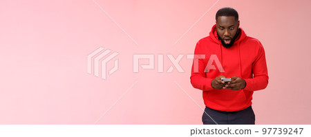 Amused focused impressed african-american bearded guy in red hoodie drop jaw widen eyes look amazed smartphone display winning hard game level answering exciting message, standing pink background Amused focused impressed african-american bearded guy in red hoodie drop jaw widen eyes look amazed smartphone display winning hard game level answering exciting message, standing pink background 97739247