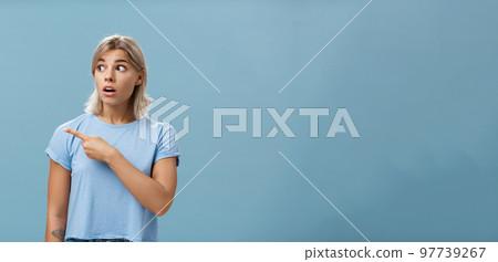 Waist-up shot of speechless shocked and impressed attractive young woman with blond hair dropping jaw staring and pointing confused and shook left standing over blue background Waist-up shot of speechless shocked and impressed attractive young woman with blond hair dropping jaw staring and pointing confused and shook left standing over blue background 97739267