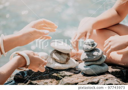 Woman with daughter bilds stones pyramid on seashore on a sunny day on the blue sea background. Happy family holidays. Pebble beach, calm sea. Concept of happy vacation on the sea, meditation, spa Woman with daughter bilds stones pyramid on seashore on a sunny day on the blue sea background. Happy family holidays. Pebble beach, calm sea. Concept of happy vacation on the sea, meditation, spa 97743973