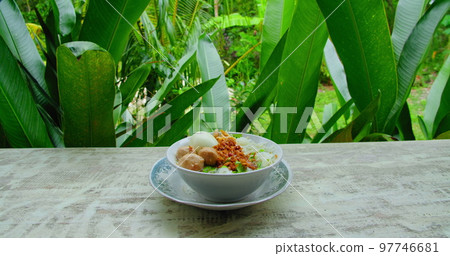 Traditional balinese food mie bakso ayam served in bowl. Plate with tasty dish of indonesian national Meatball Chicken Noodles soup. Green tropical leaves nature background. 97746681
