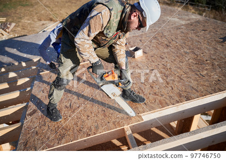 Carpenter using chainsaw for cutting wooden OSB board. Man worker building wooden frame house. Carpentry concept. 97746750