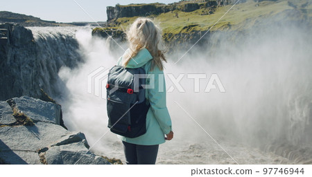 Woman standing at cliff edge looking at Detifoss waterfall in Iceland Her blond hair waving on the wind Woman standing at cliff edge looking at Detifoss waterfall in Iceland Her blond hair waving on the wind 97746944