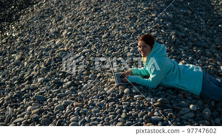 Caucasian woman lying on her stomach on a pebble beach and typing on a laptop. Remote work freelancer. Caucasian woman lying on her stomach on a pebble beach and typing on a laptop. Remote work freelancer. 97747602