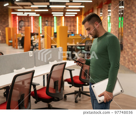 Caucasian bearded man using smartphone and holding laptop in modern coworking space.  Caucasian bearded man using smartphone and holding laptop in modern coworking space.  97748036