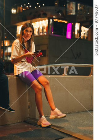 Young woman sitting with her phone on the street at night Young woman sitting with her phone on the street at night 97748037