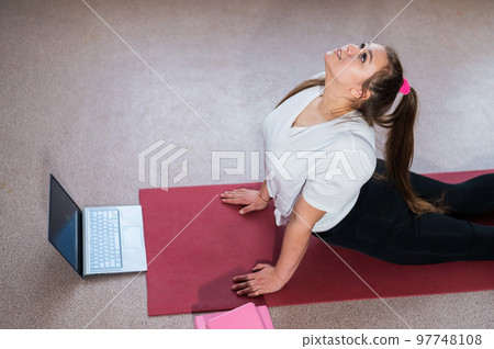 A chubby young woman is watching an online fitness lesson on a laptop. Distance sports training. A chubby young woman is watching an online fitness lesson on a laptop. Distance sports training. 97748108