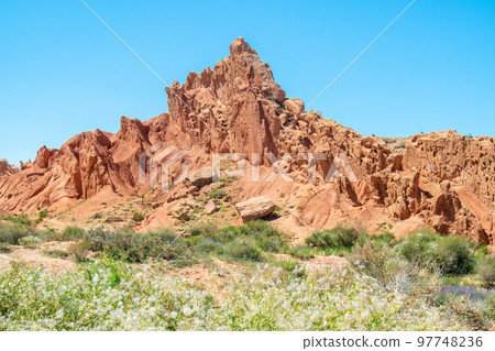 Sharp Mountain is a rock made of red orange stone and clay against a bright blue sky without clouds and desert vegetation in Fairy Tale Canyon Kyrgyzstan, Skazka Canyon. 97748236