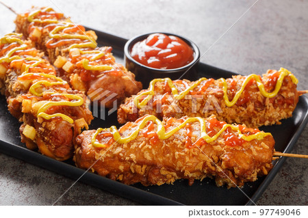 Korean corn dogs are hot dogs, cheese, French fries fried in a batter on a stick and dressed with sugar, ketchup, mustard closeup on the plate. Horizontal Korean corn dogs are hot dogs, cheese, French fries fried in a batter on a stick and dressed with sugar, ketchup, mustard closeup on the plate. Horizontal 97749046