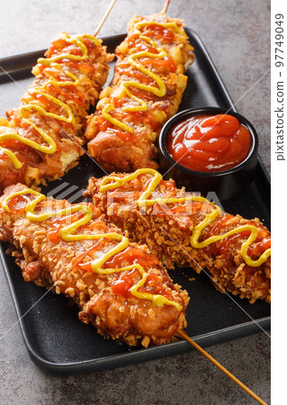 Korean corn dogs are made from sausage, mozzarella cheese on skewer and coat this in a sweet flour batter before rolling in a layer of toppings closeup on the plate. Vertical 97749049