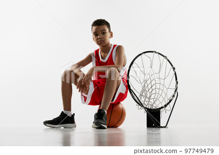 Portrait of boy in red uniform, basketball player posing with ball and basket net over grey studio background Portrait of boy in red uniform, basketball player posing with ball and basket net over grey studio background 97749479