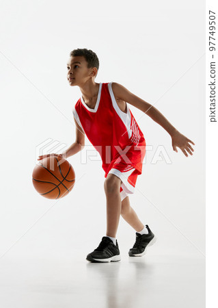 Portrait of boy in red uniform training, playing basketball over grey studio background. Dribbling exercises 97749507