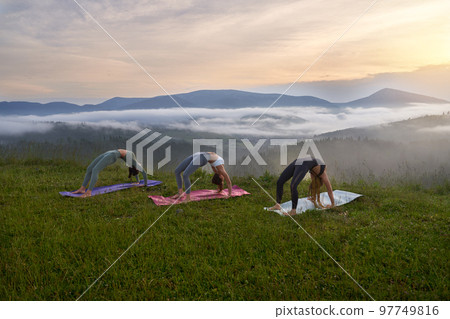 Healthy fit women in sport clothes stretching body on yoga mat among beautiful summer mountains. Three young ladies spending free time for outdoors activity.  97749816