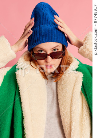 Portrait of stylish young girl in sunglasses, blue hat, green scarf and fur coat posing over pink background. Bright style Portrait of stylish young girl in sunglasses, blue hat, green scarf and fur coat posing over pink background. Bright style 97749917