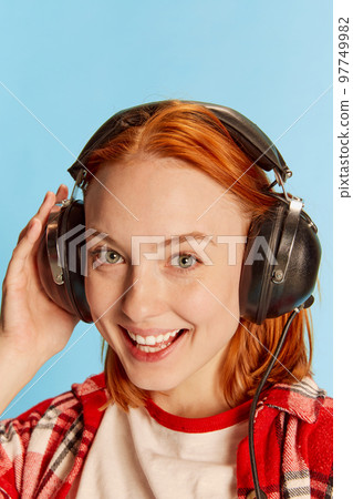 Portrait of young redhead girl in casual checkered shirt posing in headphones over blue background. Feeling happy Portrait of young redhead girl in casual checkered shirt posing in headphones over blue background. Feeling happy 97749982