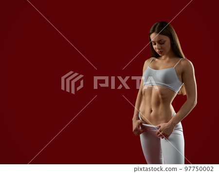 Attractive woman with press cubes in grey sport suite posing on red background, looking down. Sexy athletic fit female standing in studio. Female trainer after training. Concept of sport shape. Attractive woman with press cubes in grey sport suite posing on red background, looking down. Sexy athletic fit female standing in studio. Female trainer after training. Concept of sport shape. 97750002