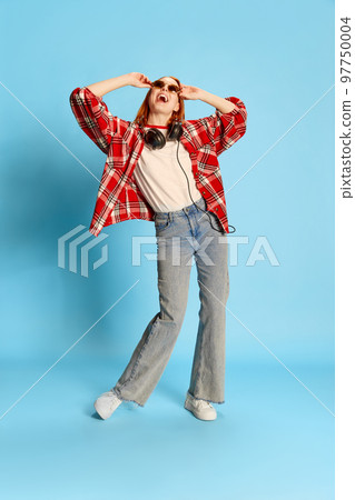 Portrait of young redhead girl in casual checkered shirt, in headphones dancing over blue background. Positivity Portrait of young redhead girl in casual checkered shirt, in headphones dancing over blue background. Positivity 97750004