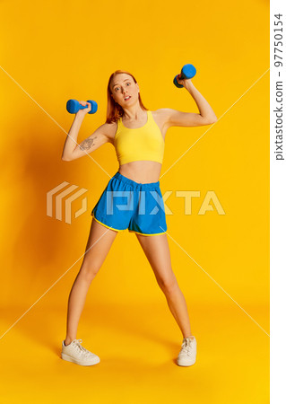 Portrait of young redhead girl in sportswear training with sports equipment over yellow background. Lifting dumbbells 97750154