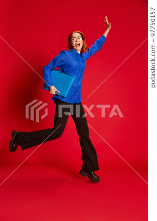 Portrait of young redhead girl in blue shirt and glasses posing in a jump with laptop over red background. Successful student 97750157