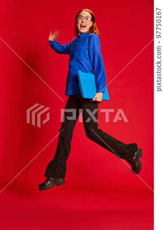 Portrait of young redhead girl in blue shirt and glasses posing in a jump with laptop over red background. Ambitions 97750167