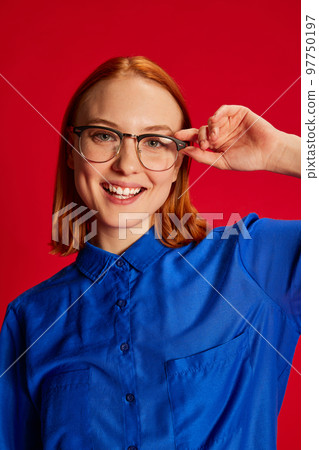 Portrait of young redhead girl in blue shirt and glasses posing, smiling over red background. Positive mood 97750197