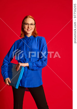 Portrait of young redhead girl in blue shirt and glasses posing with laptop over red background. Happy worker Portrait of young redhead girl in blue shirt and glasses posing with laptop over red background. Happy worker 97750205