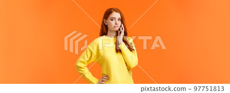 Girl catching anxiety calling someone. Perplexed or nervous cute redhead woman biting lip and looking away as having conversation on mobile phone, standing orange background sad 97751813