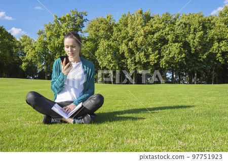 A young woman reading a message on the phone in the park on the grass. Education and knowledge A young woman reading a message on the phone in the park on the grass. Education and knowledge 97751923