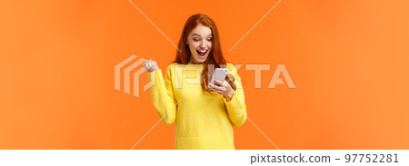 Girl received thousand likes under new picture. Cheerful and triumphing pretty redhead female fist pump say yes, cheering or celebrating good news, looking smartphone screen, orange background 97752281