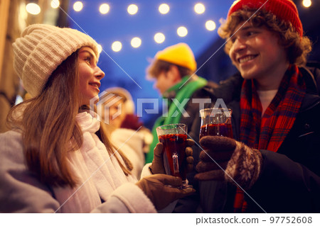 Happy young men and girls, friends walking at festive Christmas street and drinking mulled wine. Concept of holidays, winter, New Years fairs, friendship Happy young men and girls, friends walking at festive Christmas street and drinking mulled wine. Concept of holidays, winter, New Years fairs, friendship 97752608