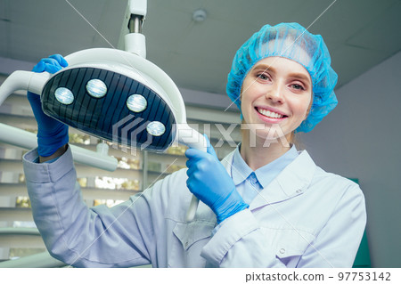 Portrait of smiling female surgeon in surgical uniform at operating room posing near operational light in laboratory Portrait of smiling female surgeon in surgical uniform at operating room posing near operational light in laboratory 97753142