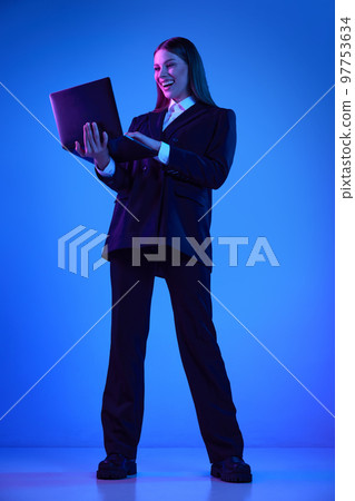 Woman in stylish official suit working on laptop over blue background in neon light. Successful cooperation 97753634