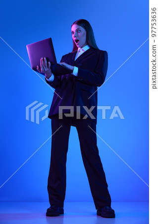Woman in stylish official suit working on laptop over blue background in neon light. Shocked employee 97753636
