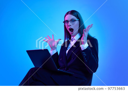 Woman in stylish official suit working on laptop over blue background in neon light. Shocked business woman 97753663