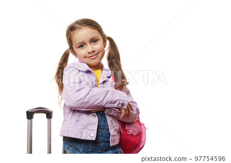 Caucasian preschooler lovely little traveler girl with two ponytails, dressed in purple jacket and blue denim overalls, smiling at camera, posing with pink backpack and suitcase on white background. 97754596