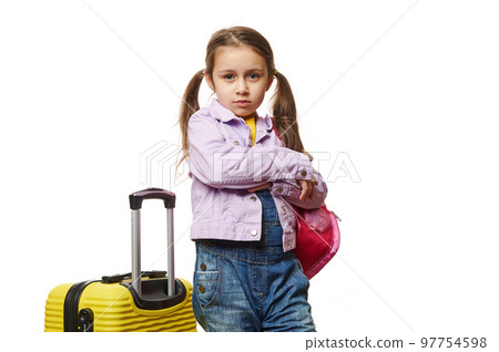 Isolated portrait on white background of a little child girl with two ponytails, dressed in purple jacket, posing with her arms crossed, standing near yellow suitcase over white background. Copy Space 97754598