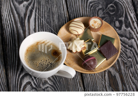 A cup of black coffee in a white cup. Chocolate in foil and chocolates. On pine boards. 97756228