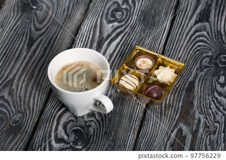 A cup of black coffee in a white cup. Chocolate in foil and chocolates. On pine boards. 97756229