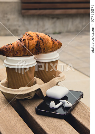 Fresh baked chocolate croissant on Two paper cups with lid for tea to go. Breakfast Coffee take away on the table. Take-out coffees with brown paper cup holder. Mobile phone with wireless headphones. Fresh baked chocolate croissant on Two paper cups with lid for tea to go. Breakfast Coffee take away on the table. Take-out coffees with brown paper cup holder. Mobile phone with wireless headphones. 97756875