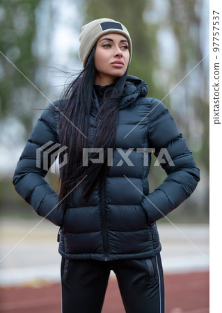 Beautiful brunette woman posing at the sport stadium in jacket and hat Beautiful brunette woman posing at the sport stadium in jacket and hat 97757537