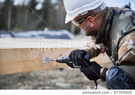 Man building wooden frame house. Close up of male worker drilling hole by electric drill in timber framing. Builder wearing white helmet and protective goggles. Carpentry concept. 97769008