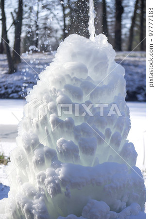 Iced fountain in the park on a winter sunny day. Fountain with frozen water. A jet of water from a fountain that iced over in the cold. Background covered with ice and snow in the park on day Iced fountain in the park on a winter sunny day. Fountain with frozen water. A jet of water from a fountain that iced over in the cold. Background covered with ice and snow in the park on day 97777183