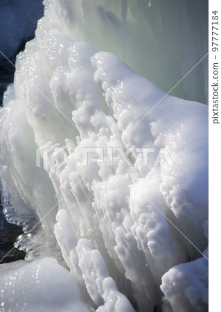 Iced fountain in the park on a winter sunny day. Fountain with frozen water. A jet of water from a fountain that iced over in the cold. Background covered with ice and snow in the park on day 97777184